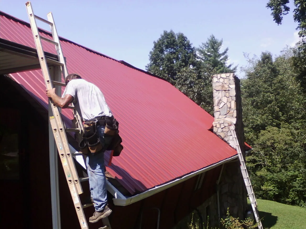 Roofer on a ladder beside a red metal roof and stone chimney — Soffit & Fascia Repair by Roofing Brothers in Thorntown